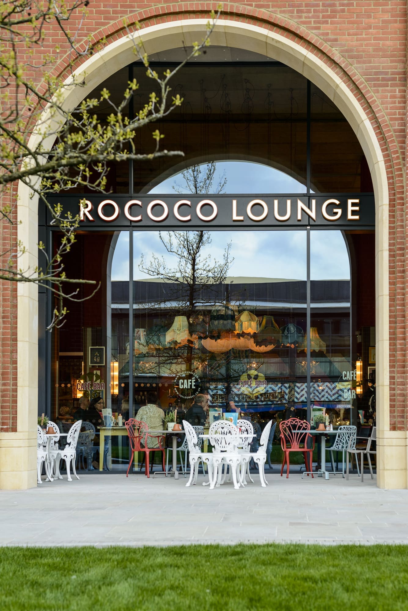 A large arched window with the sign "Rococo Lounge" above it reveals a stylish Rococo-inspired café interior. Outside, white and red metal chairs and tables sit on a patio, framed by a tree and green grass in the foreground.