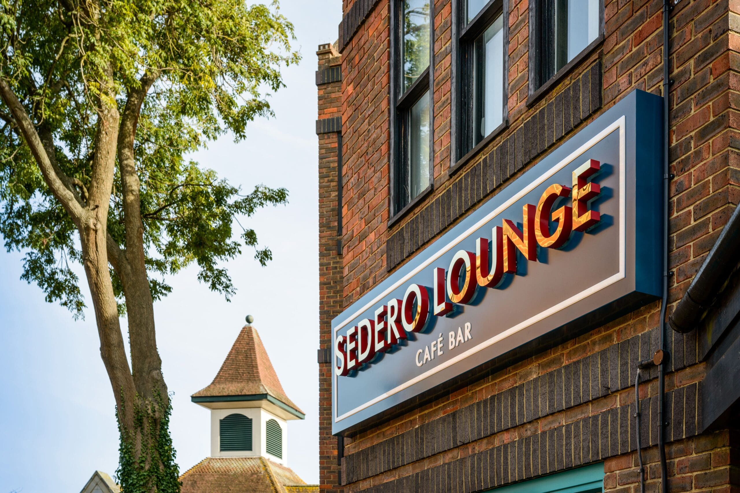 The photo shows the Sedero Lounge Café Bar, a brick building with its sign visible. In the background, a leafy tree and another building with a pointed roof and louvered windows stand under a clear sky.