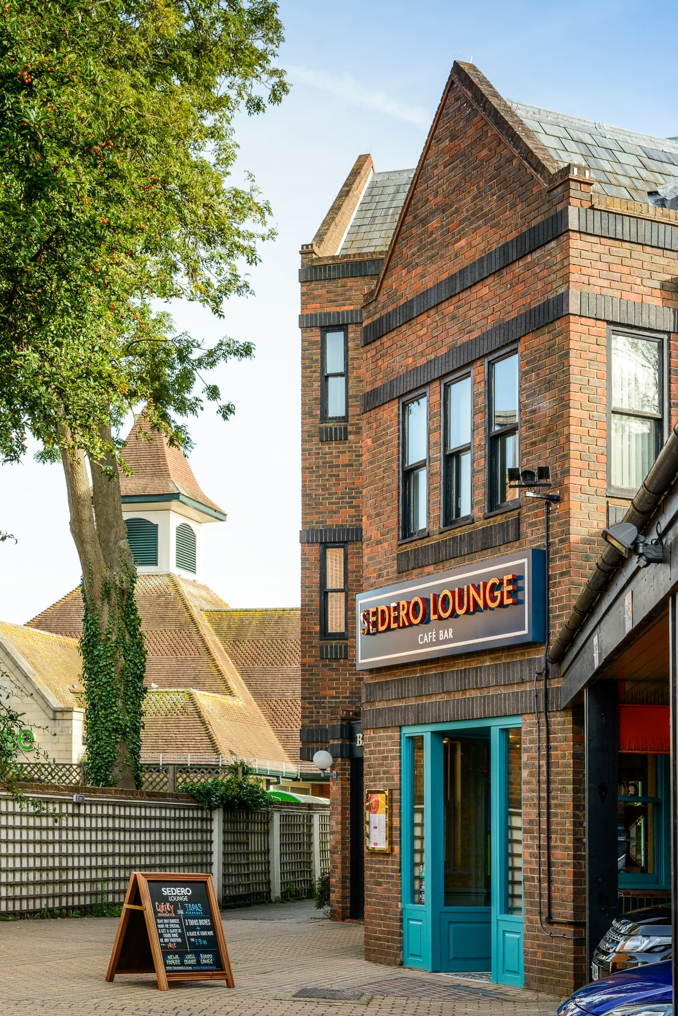 A brick building with a sign reading "Sero Lounge Cafe Bar" above blue doors. An outdoor menu board stands nearby. In the background, the Sedero area features other buildings and a tree under a clear sky.