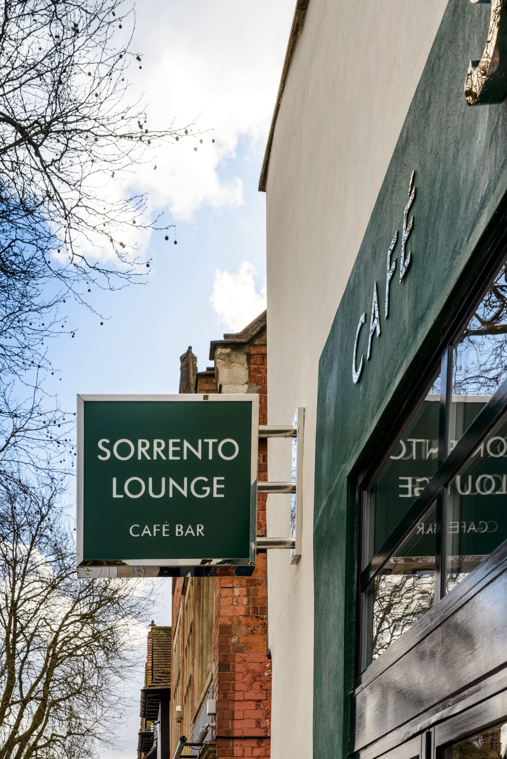 A green sign reading "SORRENTO LOUNGE CAFÉ BAR" hangs outside a Sorrento building with a matching green wall featuring "CAFÉ." Leafless trees and a partly cloudy sky are in the background.