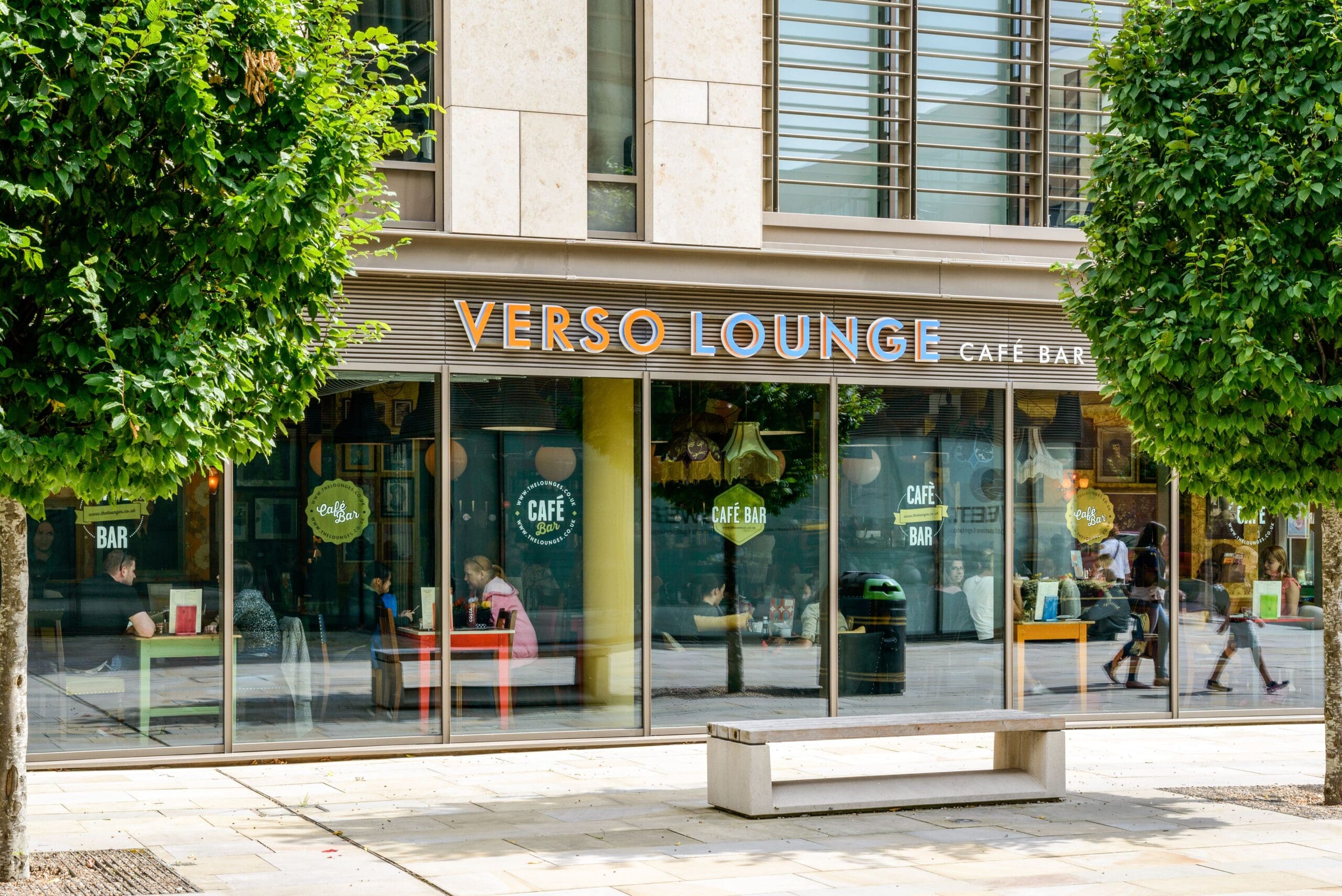 Street view of Verso Lounge Café Bar with large glass windows, people relaxing inside, and trees framing the entrance. A bus shelter is visible on the pavement in front of Verso, enhancing its inviting urban vibe.