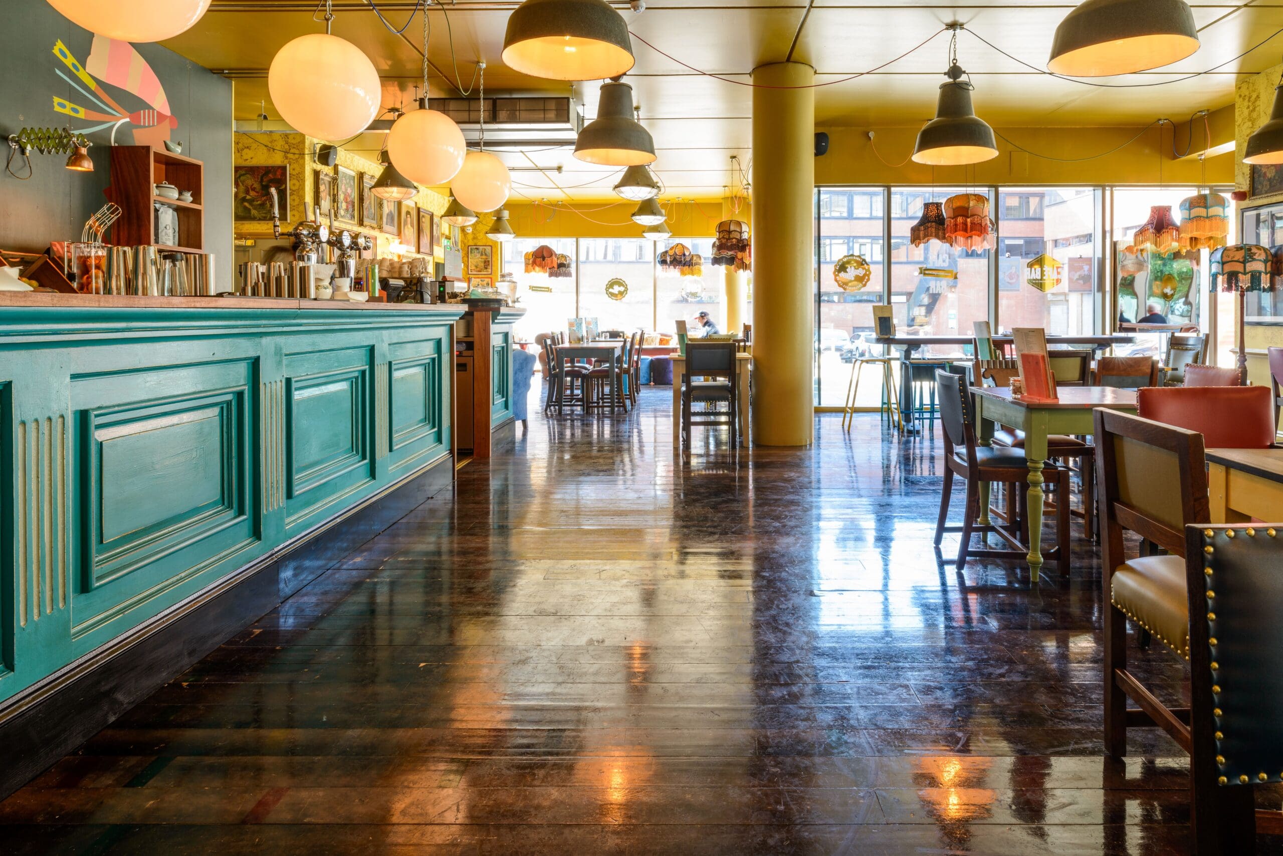 A brightly lit, modern café with a teal counter, wooden floors, colorful chairs, and Verso-inspired décor. Round hanging lights and large windows fill the cozy space with natural light, while tables are neatly arranged for an inviting atmosphere.