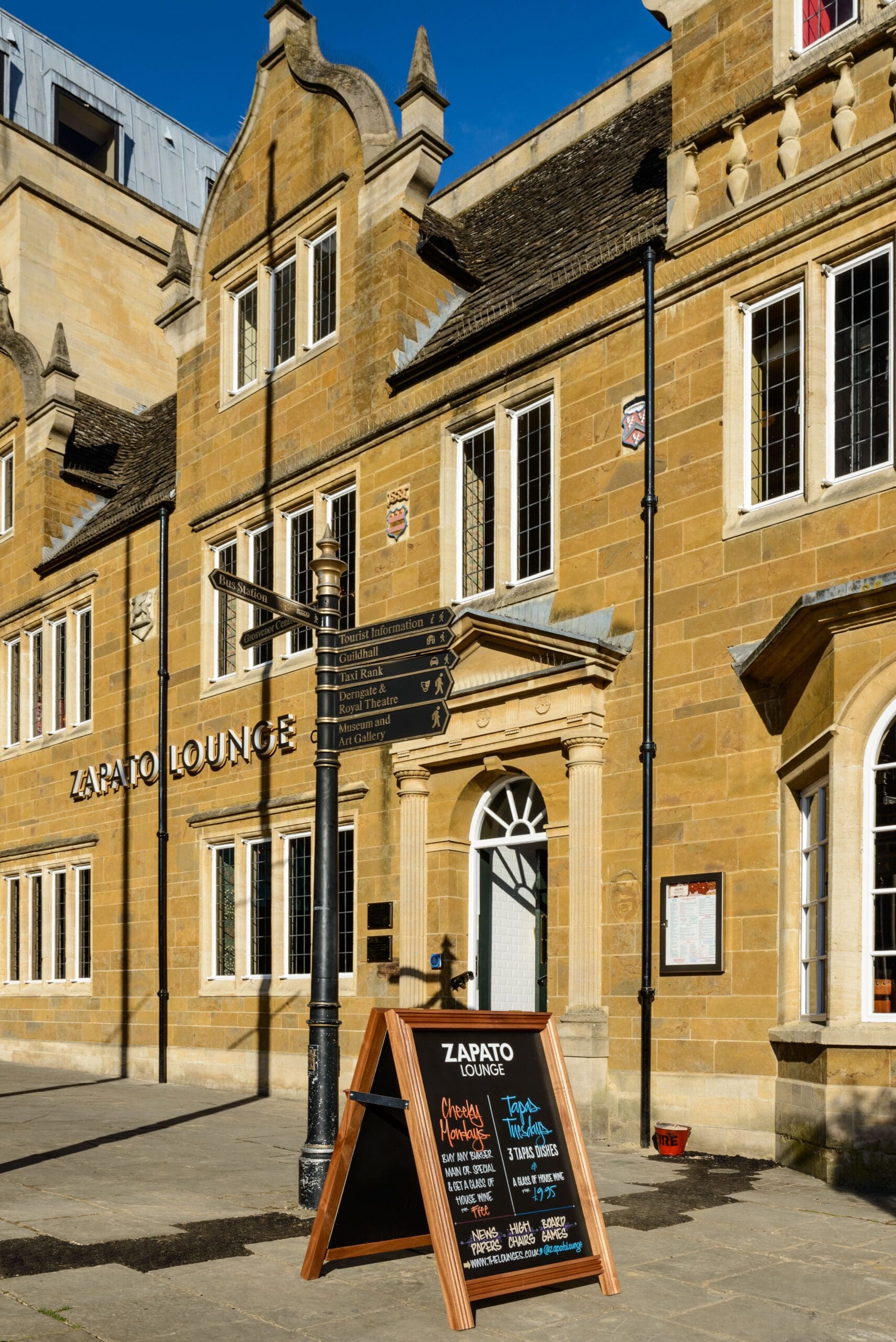 A sandwich board with a Zapato Lounge menu stands outside the historic yellow stone building on a sunny day, with a black street signpost and blue sky in the background.
