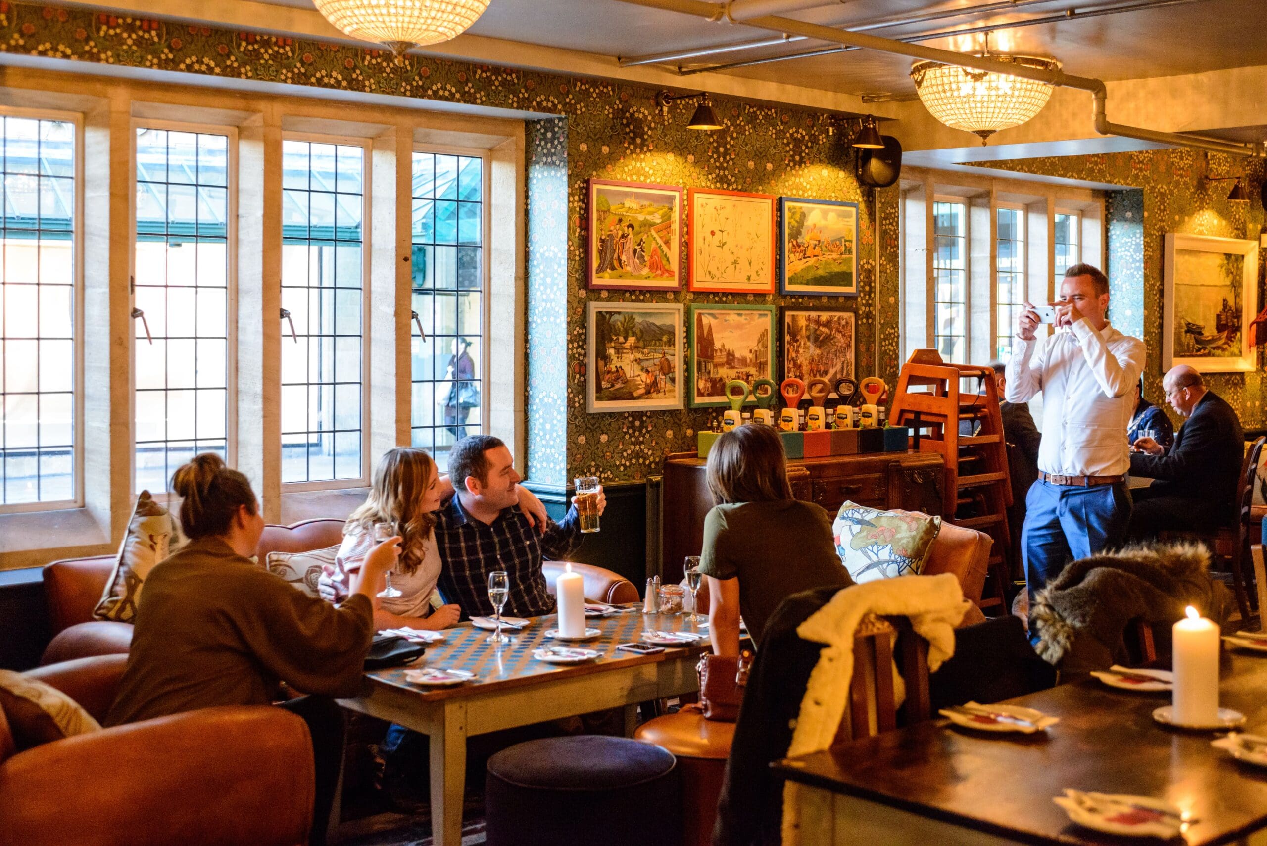 A cozy pub interior with people sitting at tables, chatting and drinking. One man in Zapato shoes stands taking a photo with his phone, while another drinks near the window. Warm lighting and framed pictures decorate the walls.