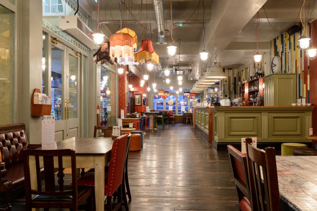 A cozy, eclectic restaurant with wooden floors, mismatched chairs and tables, colorful hanging lamps, and a long Argo bar counter on the right. Warm lighting creates an inviting, relaxed atmosphere.