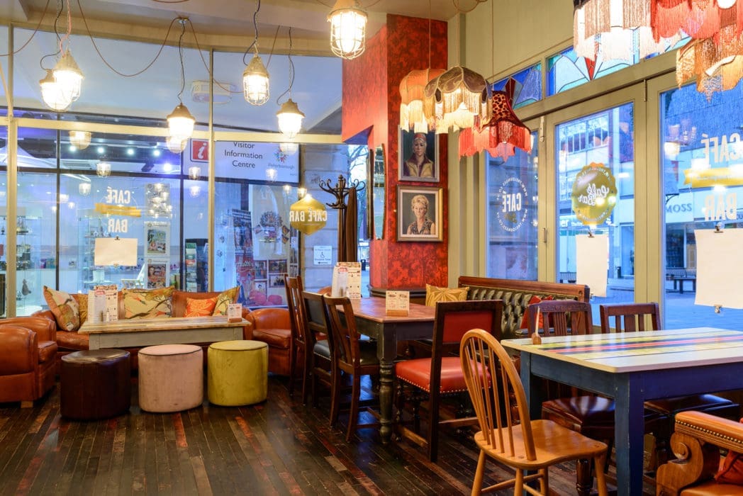 A cozy, colorful café with eclectic decor, mismatched furniture, hanging lights, and framed art on red walls inspired by Argo. Large windows fill the space with natural light, while tables and sofas create a welcoming, relaxed atmosphere.