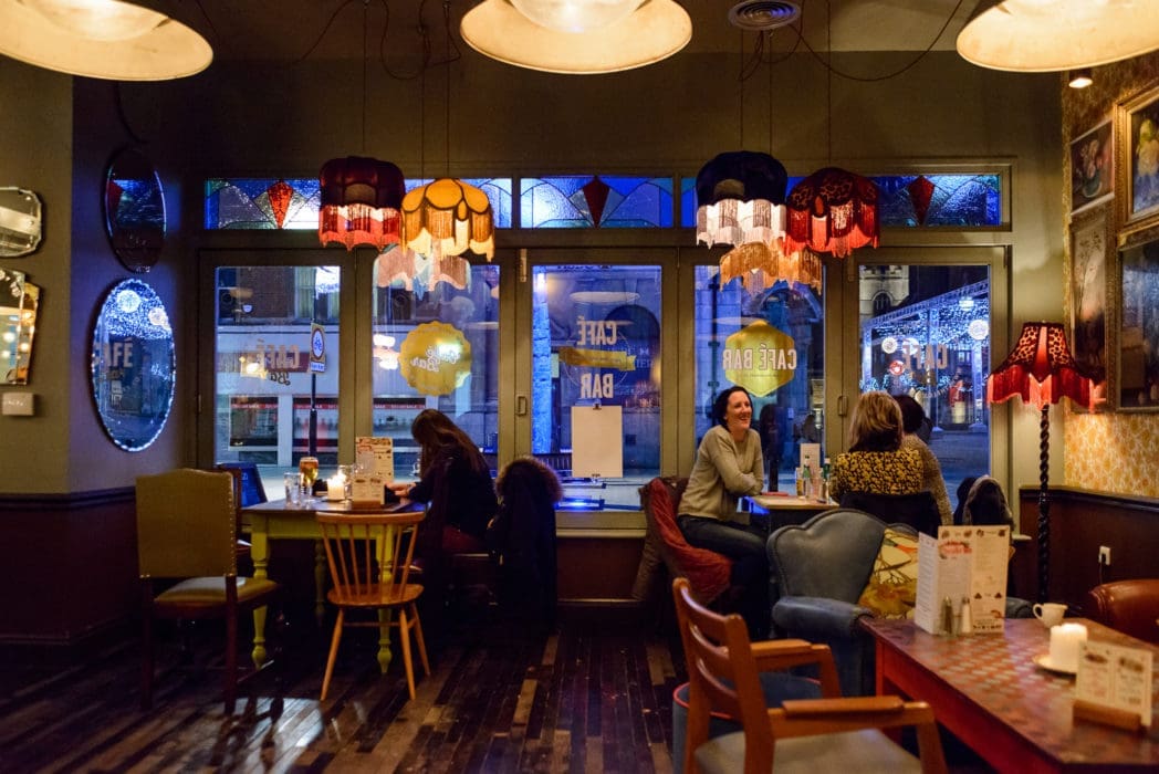 Cozy Argo café interior with vintage lamps, colorful chairs, and groups of people chatting at tables. Large windows reveal streetlights and the nighttime city outside. Warm, inviting atmosphere with eclectic decor.
