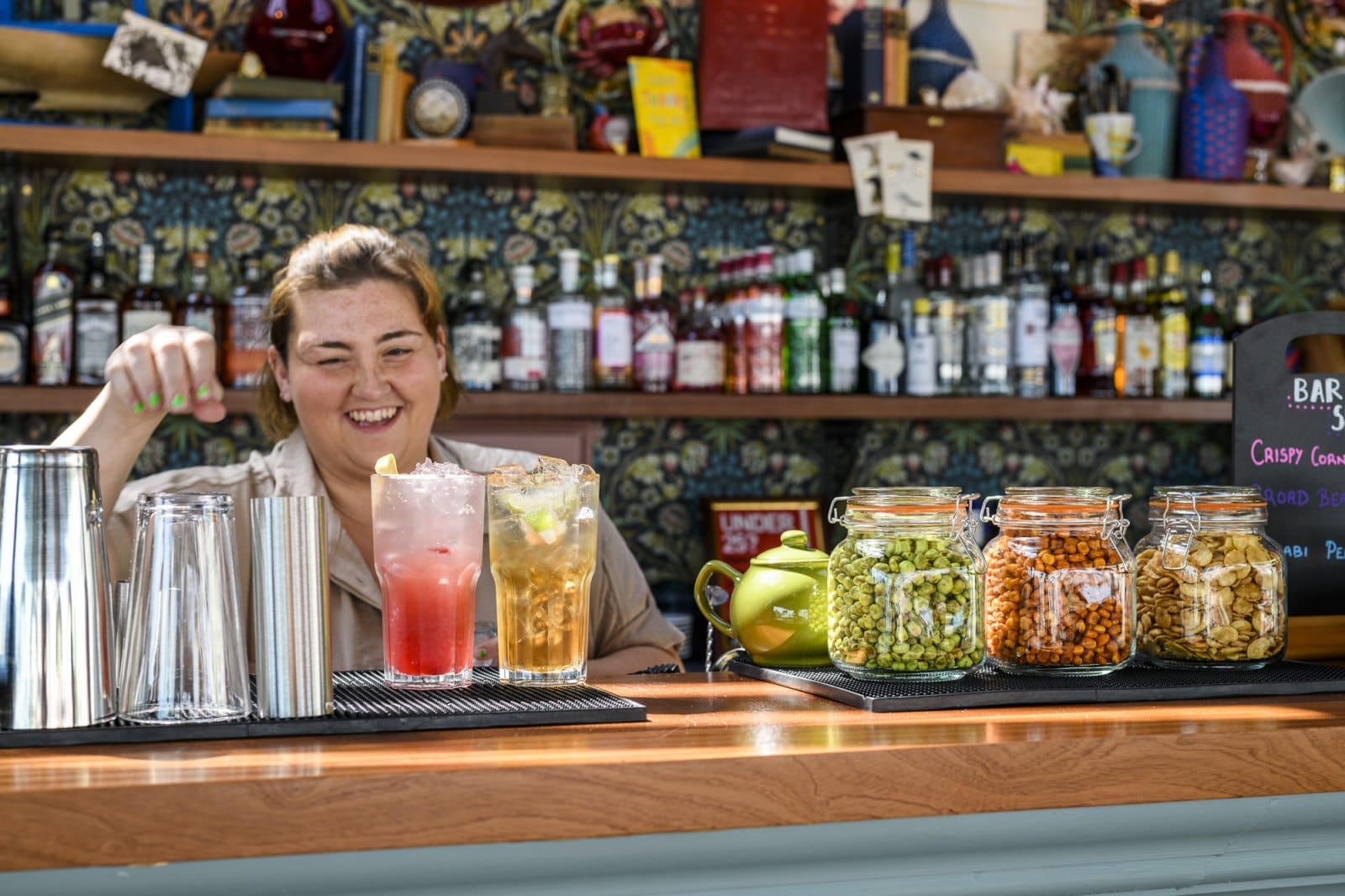 A smiling bartender stands behind a bar with two colorful drinks, jars of snacks, and cocktail shakers; shelves of bottles and Bianco spirits mingle with decorative items in the background.