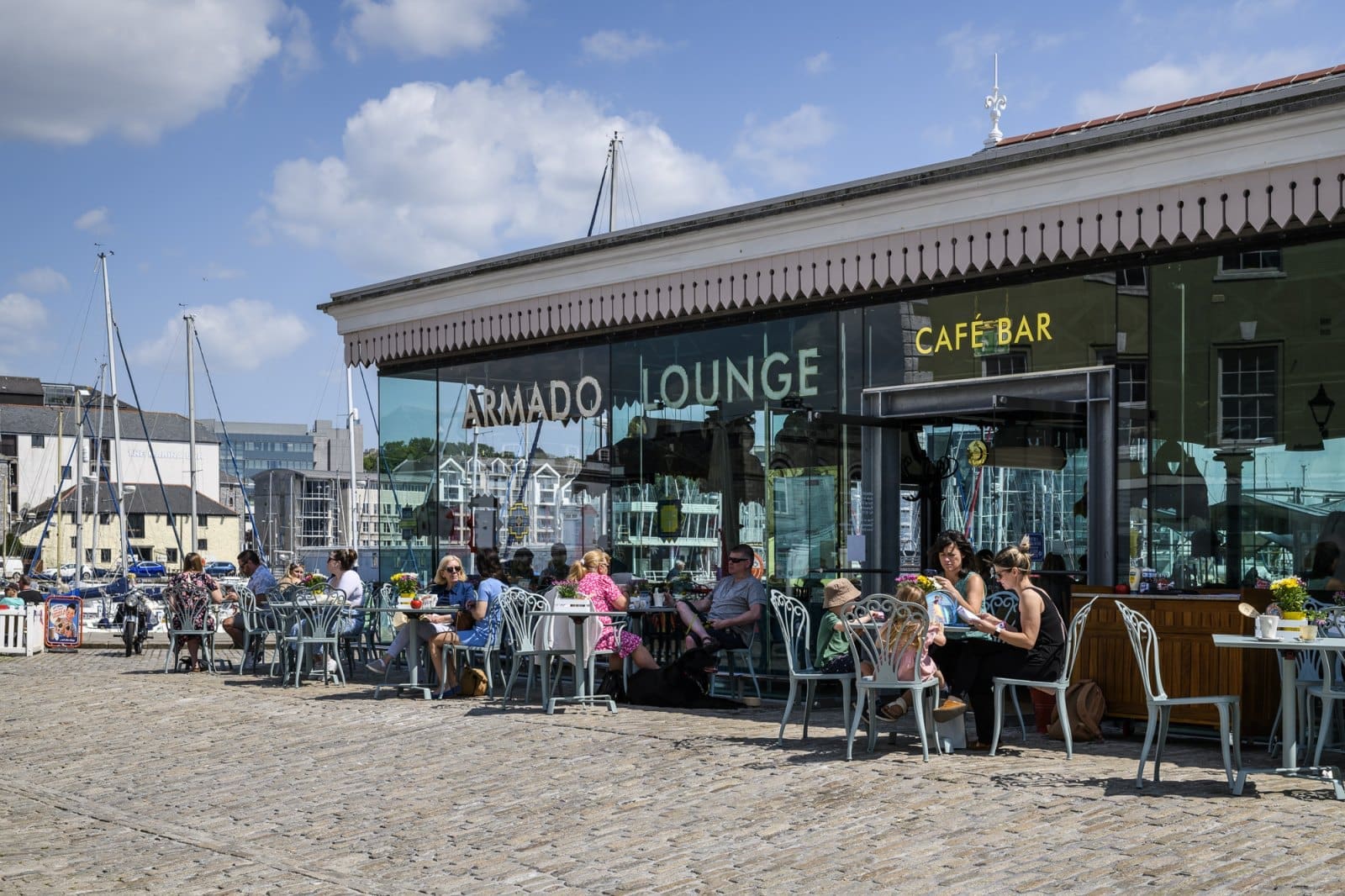 People sit at outdoor tables in front of Armado Lounge Café Bar on a sunny day, sipping Bianco and chatting, with boats and buildings in the background. Some enjoy a meal or drinks, soaking up the lively harbor atmosphere.