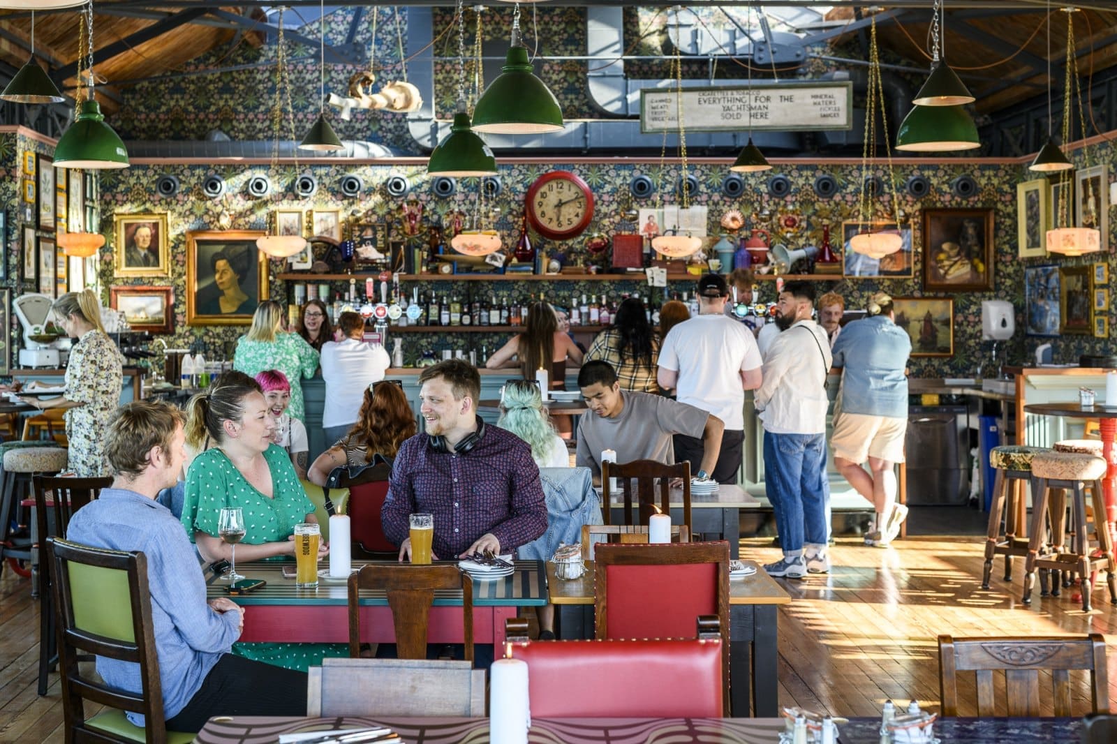 People dining and chatting at lively, eclectic Bianco restaurant with colorful decor, framed art, a clock, and a busy bar area where several people are ordering drinks. Sunlight streams in through large windows.