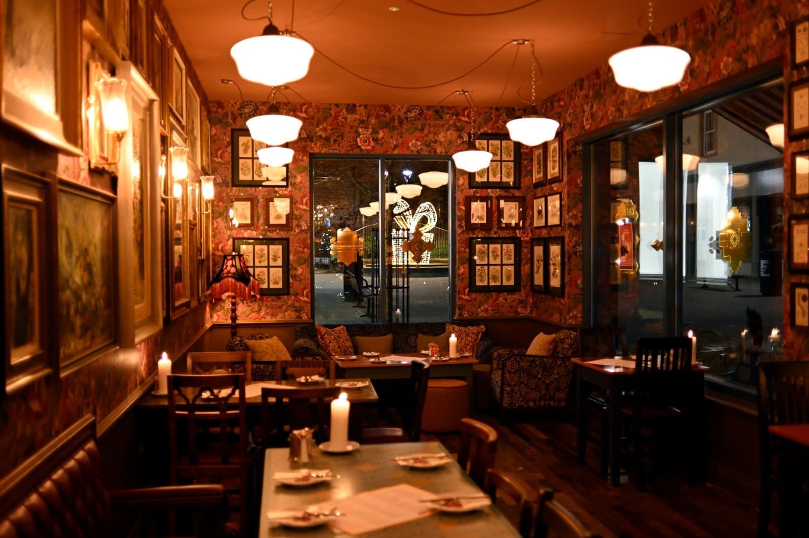 A warmly lit, cozy restaurant—Arturo—with vintage decor, framed pictures on patterned walls, candles on tables, and large windows revealing a softly illuminated street outside at night.