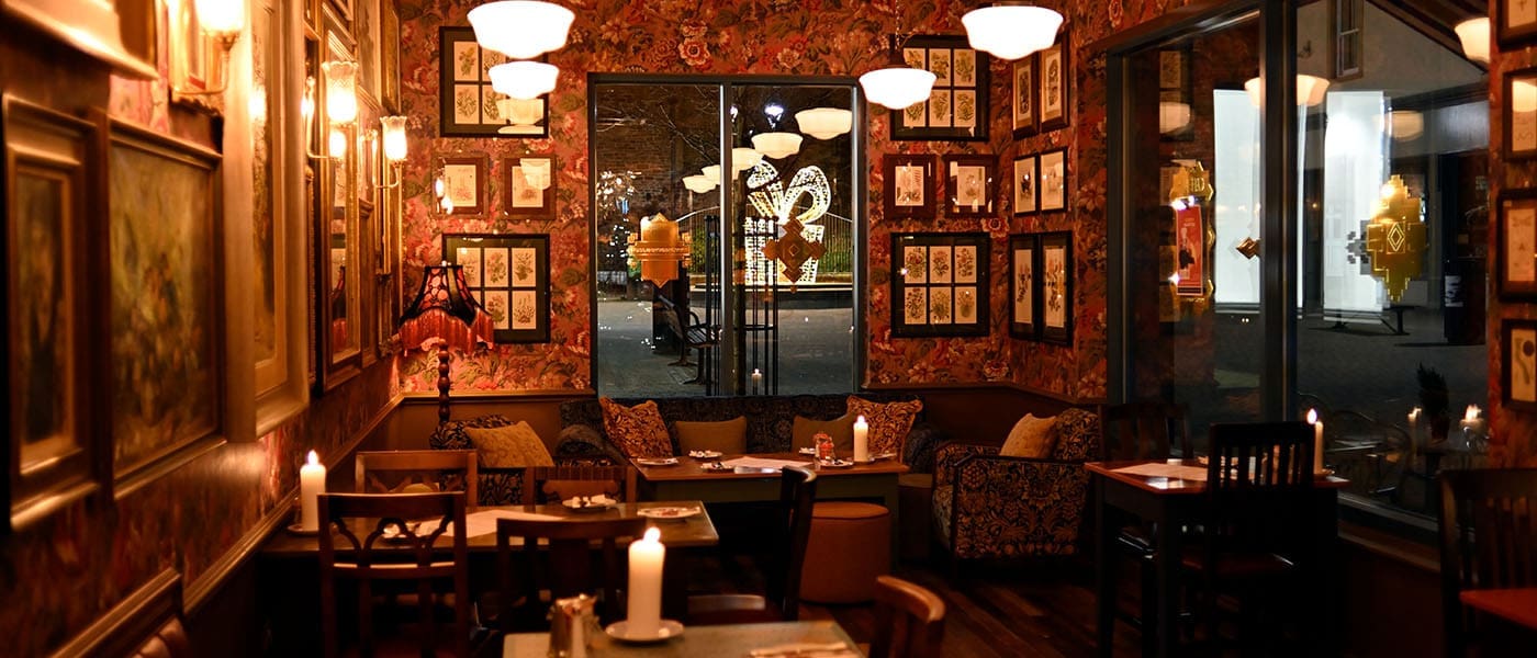 A cozy, dimly lit restaurant with floral wallpaper, framed pictures on the walls, wooden tables and chairs, and glowing candles on each table. Large windows reveal Arturo’s softly illuminated street outside.