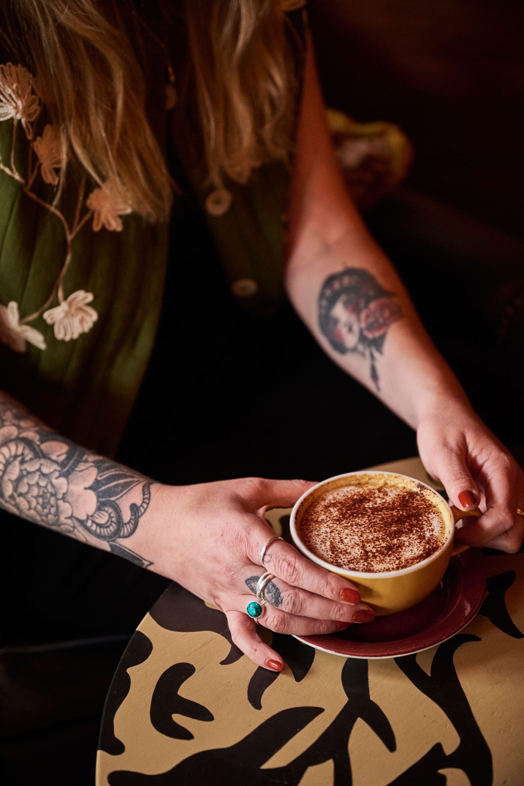 A person with long hair and tattooed arms enjoys a cup of cappuccino topped with frothy milk and cinnamon on a decorative table, savoring a relaxing tiempo. They wear rings and an embroidered garment, adding to the scene’s unique charm.