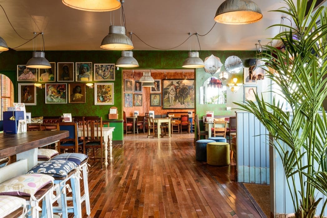 A cozy, eclectic café with wooden floors, a mix of colorful chairs and stools, green walls adorned with various framed artworks, large hanging lights, and leafy plants adding warmth to the inviting Bacco space.