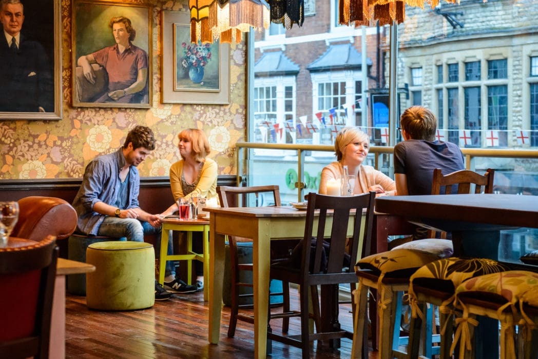 People sit at tables in a cozy, colorful café with floral wallpaper and large windows. Two pairs chat and smile at Bacco, while street decorations are visible outside. The atmosphere is warm and inviting.