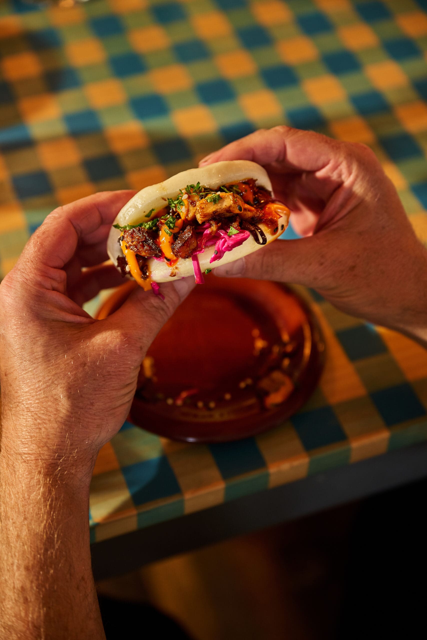 A person holds a steamed bao bun filled with colorful vegetables, meat, and sauce over a table with a checkered tablecloth.