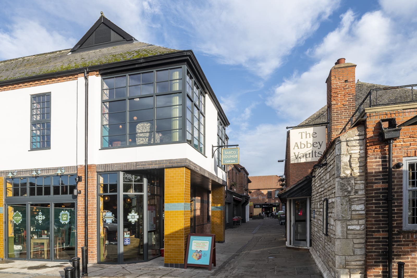 A modern building with large windows and yellow tiles stands beside a brick building labeled "The Abbey Vaults" Barco on a sunny day, with a narrow alley running between them and a bright blue sky above.