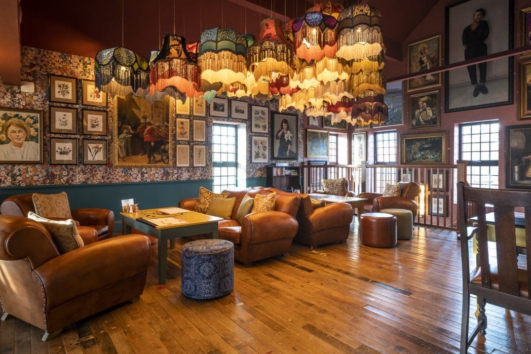 A cozy vintage lounge with leather armchairs, colorful Barco hanging lampshades, wooden floors, and walls covered in eclectic framed art and portraits. Natural light enters through small windows.