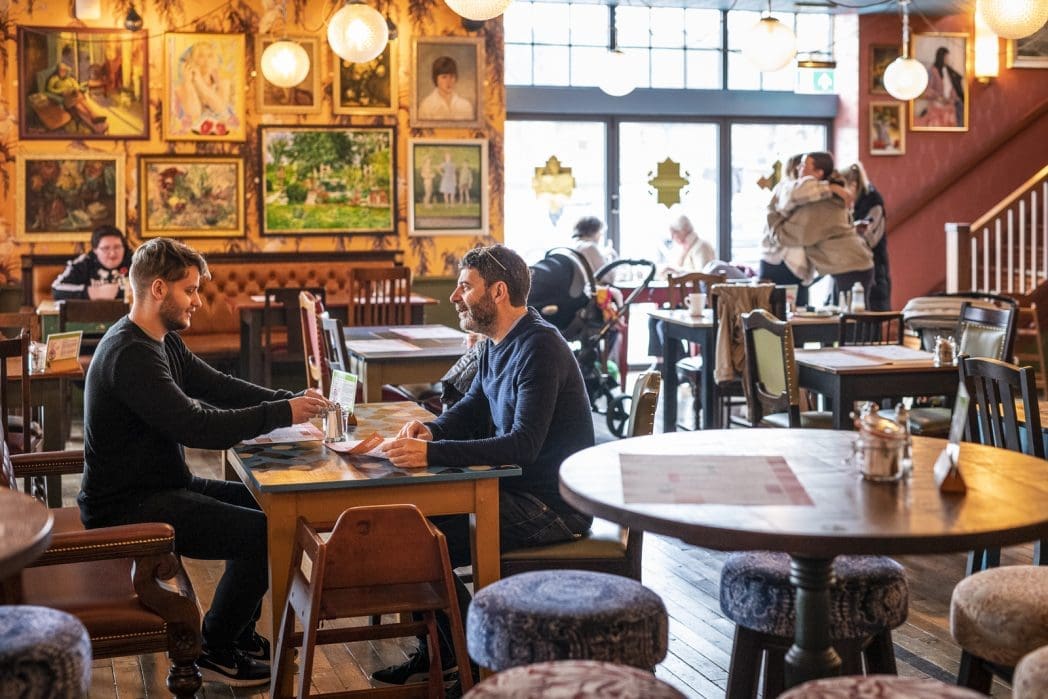 Two men sit and talk at a table in a cozy, warmly lit café filled with eclectic furniture, Barco decor, and artwork on the walls. Other patrons are visible in the background, creating a lively, welcoming atmosphere.