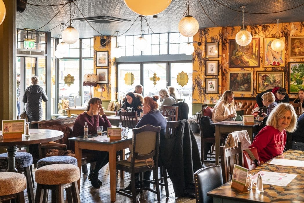 A cozy, brightly lit café with people dining at wooden tables—some linger near the Barco espresso machine. Framed art decorates yellow walls, large windows let in natural light, and diners are engaged in conversation as a few people enter the café.