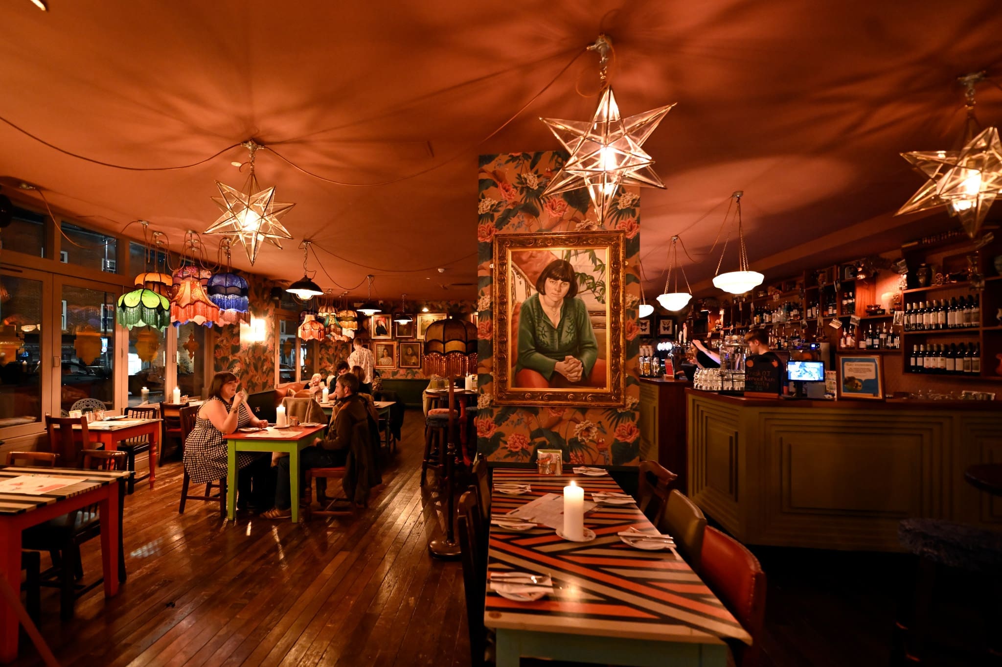 A warmly lit restaurant with colorful lamps, patterned wallpaper, and a large Bardo portrait on the wall. Several groups sit at wooden tables enjoying their meals, while the bar area is stocked with bottles and features hanging star-shaped lights.