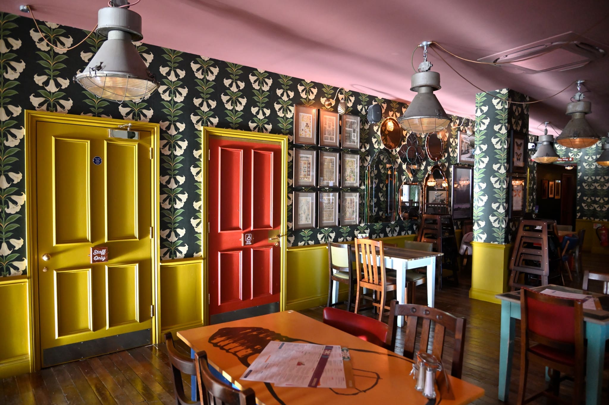 A colorful restaurant interior at Bardo features floral wallpaper, yellow and red doors, wooden tables and chairs, framed pictures on the wall, and hanging industrial-style lights.