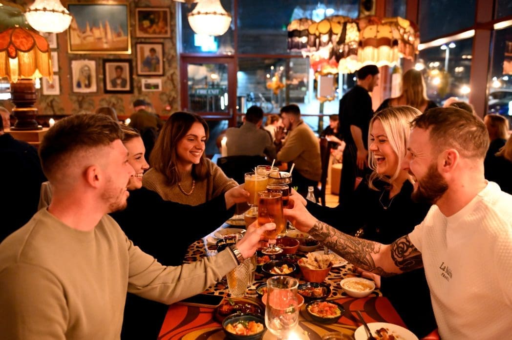 A group of five friends sit around a table in a cozy, warmly lit Barranco restaurant, smiling and raising their glasses for a toast. The table is filled with food, and other diners are visible in the background.