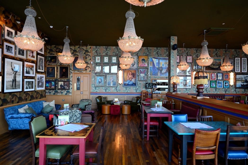 A vibrant, eclectic Barranco restaurant with wooden floors, chandeliers, mismatched chairs and sofas, and walls covered in framed art and photographs. The room feels cozy and artistic with colorful tables and soft lighting.