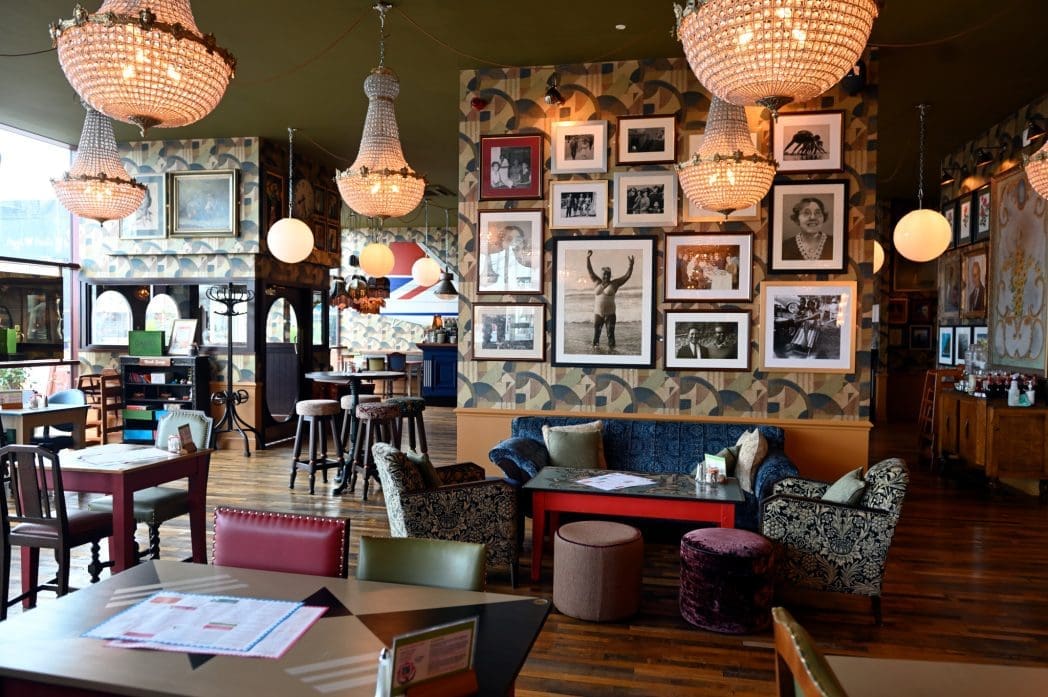 A cozy Barranco restaurant interior with eclectic furniture, chandeliers, wooden floors, and framed black-and-white photos on patterned walls. Tables and chairs are arranged throughout the space, creating a warm, inviting atmosphere.