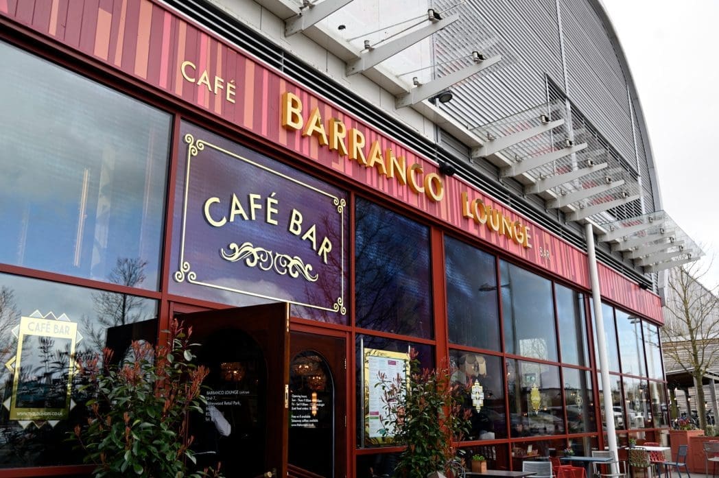 Exterior view of Barranco Lounge, a stylish café bar with large maroon and gold Barranco signage, glass windows reflecting trees, and leafy plants welcoming guests at the entrance.