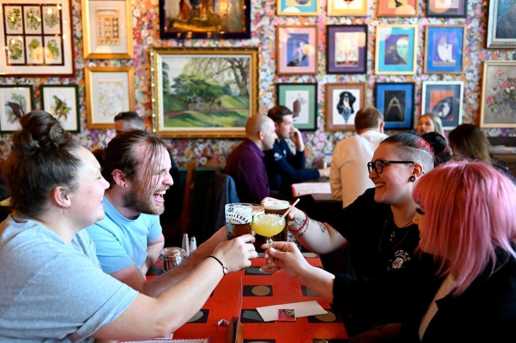 Four people sit at a lively, colorful bar or restaurant, clinking their Bianco cocktails and smiling. The walls behind them are decorated with framed pictures and artwork, while other groups enjoy the vibrant atmosphere in the background.