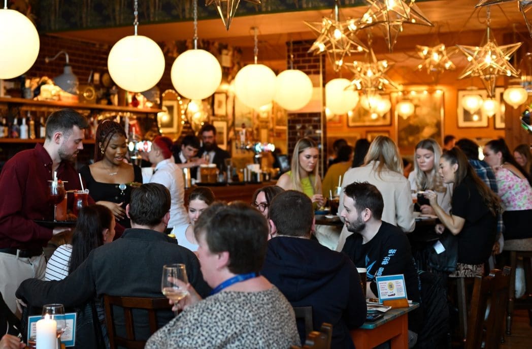A lively restaurant scene with people sitting at tables and the bar, talking and enjoying Bianco drinks. Warm lighting from hanging globe and star-shaped lamps creates a cozy atmosphere as servers move between tables.