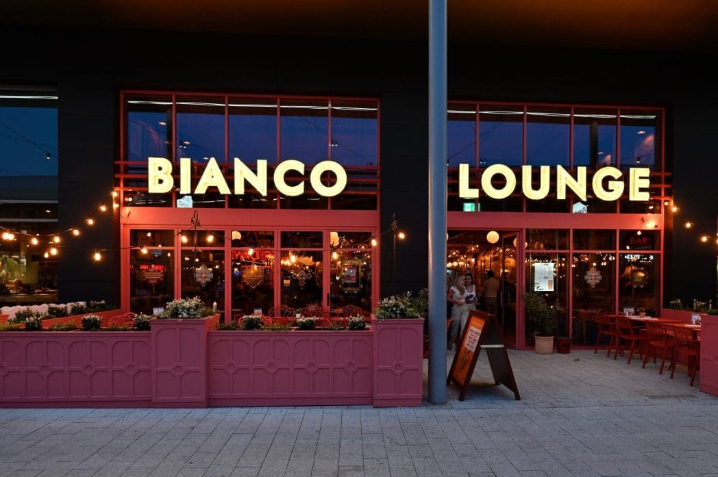 The exterior of the modern Bianco Lounge glows at dusk, with large illuminated signs, red-painted window frames, string lights, outdoor seating, and planter boxes filled with flowers.