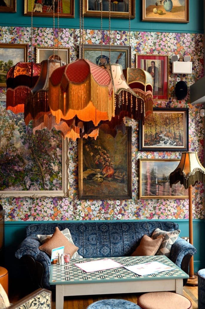A cozy vintage room with patterned wallpaper, eclectic framed artwork, a blue Bianco sofa with cushions, a wooden table, and ornate hanging lamps with fringed shades. A floor lamp with a fringed shade stands in the corner.