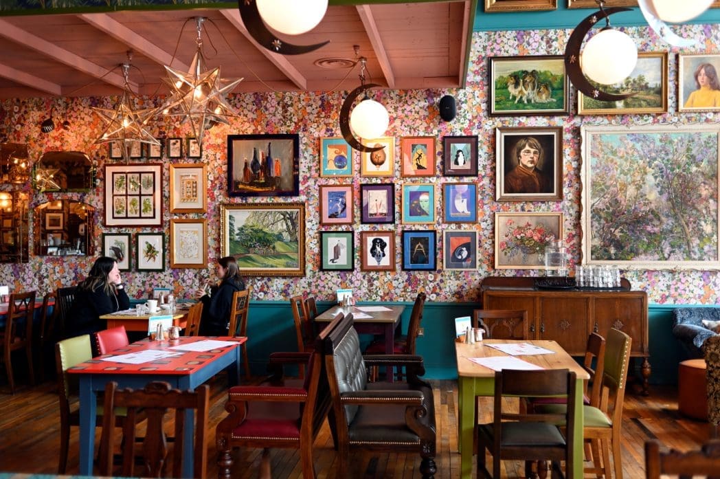 A colorful cafe with Bianco floral wallpaper, eclectic artwork and portraits covering the walls, wooden tables and chairs, and two people sitting and talking at a table. The atmosphere is vibrant and cozy.