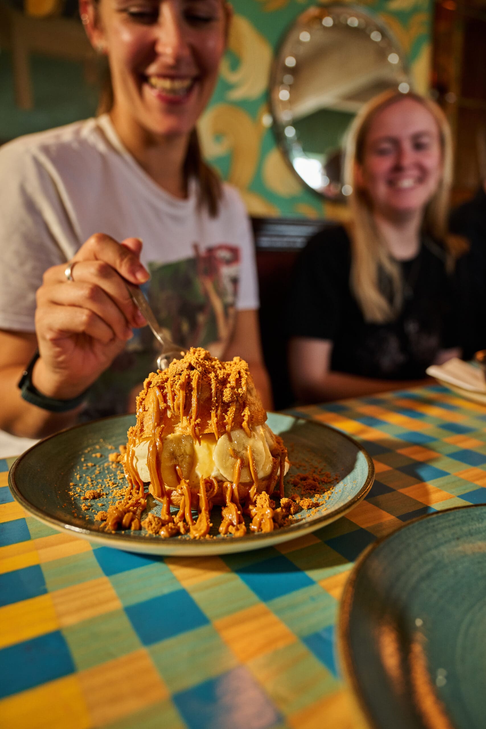 Two smiling women sit at a colorful checkered table as one prepares to eat a dessert topped with caramel sauce and crumbled cookies, presented on a teal plate.