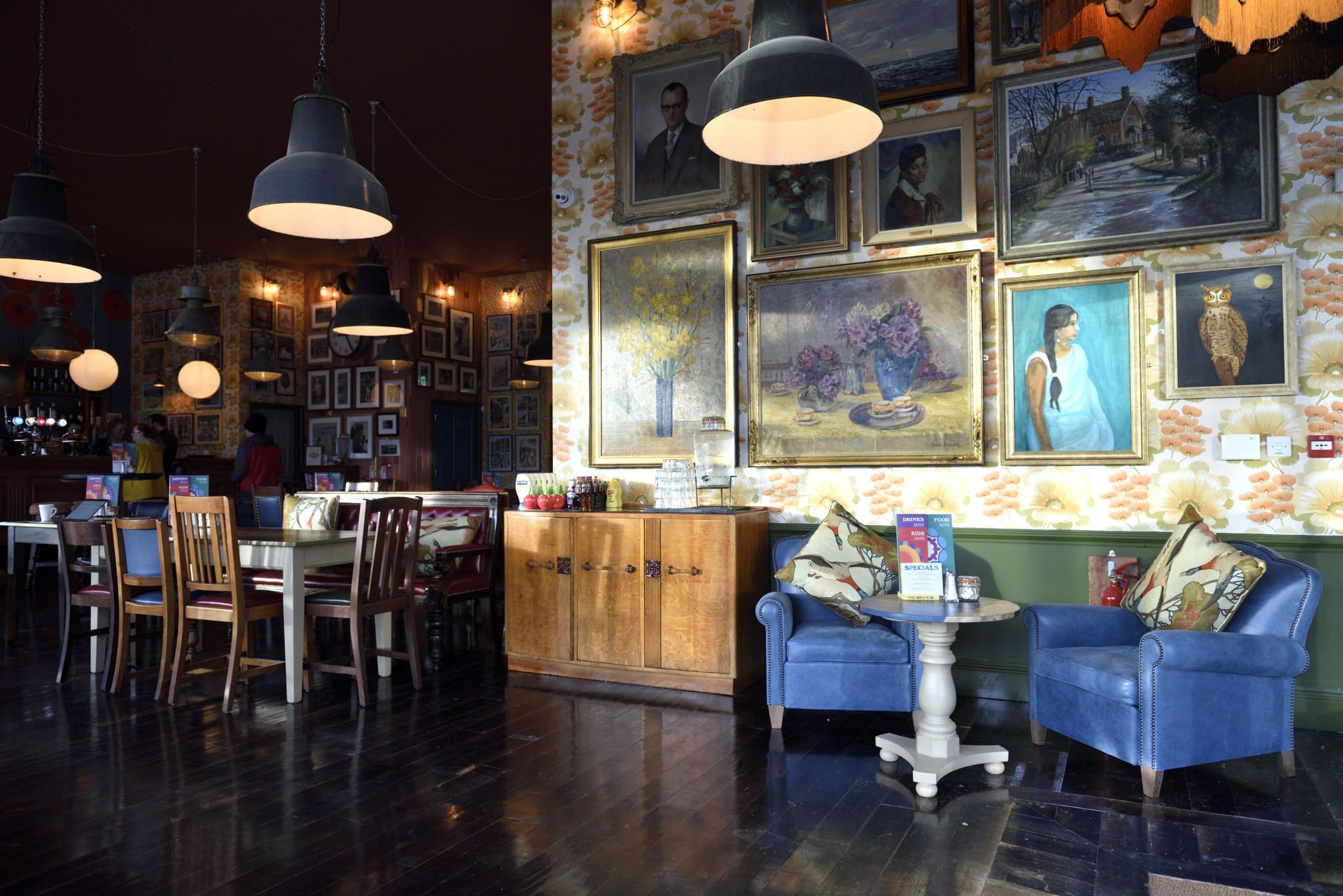 A cozy, eclectic lounge with dark wood floors, blue armchairs, a cabinet, and tables. The walls are covered with paintings, calico floral wallpaper, and large hanging lamps provide warm light.