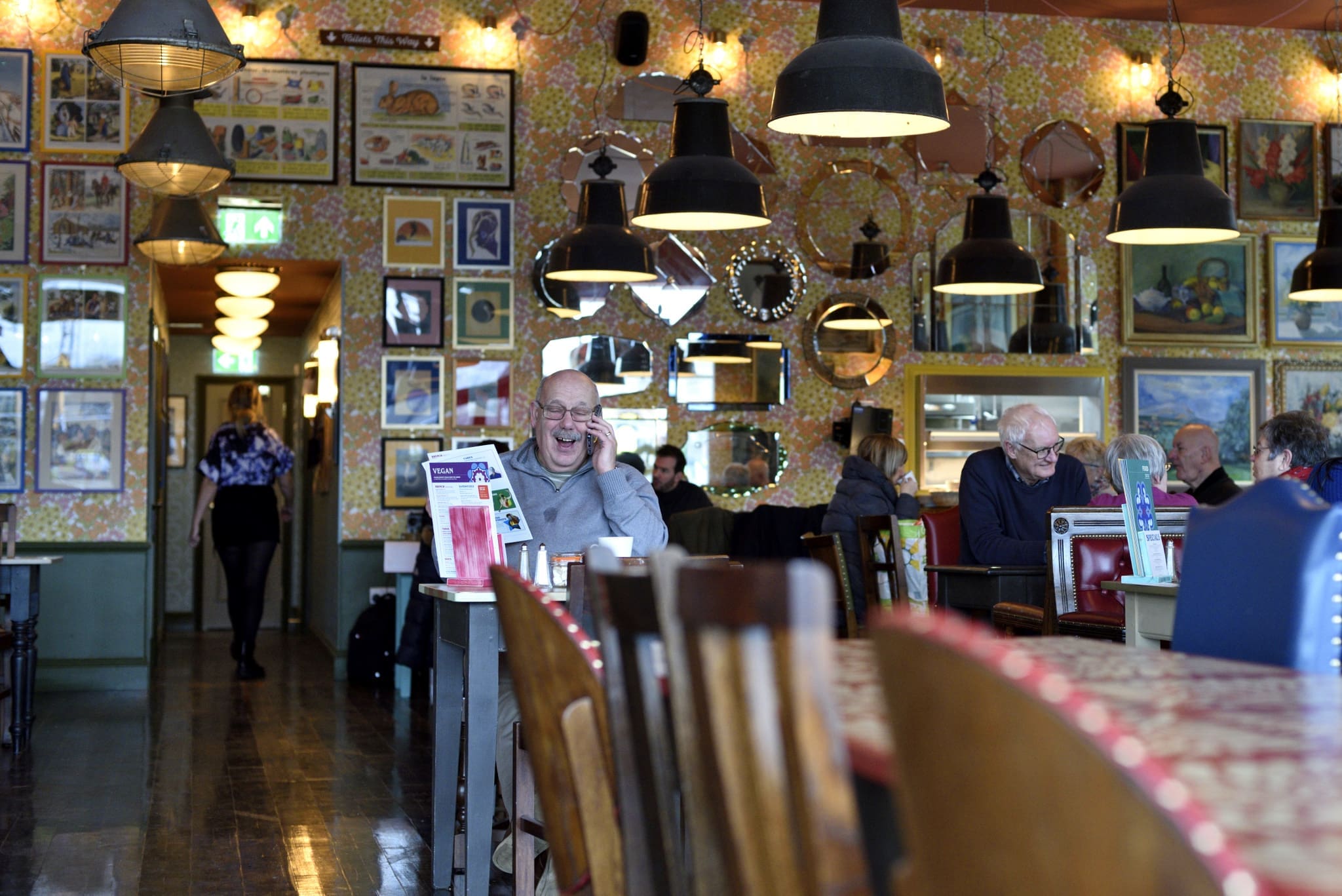 A man sits at a table in a lively, calico-colored restaurant, smiling and holding a phone to his ear. Other diners are present, and the walls are covered with framed pictures and mirrors. Warm lighting fills the room.