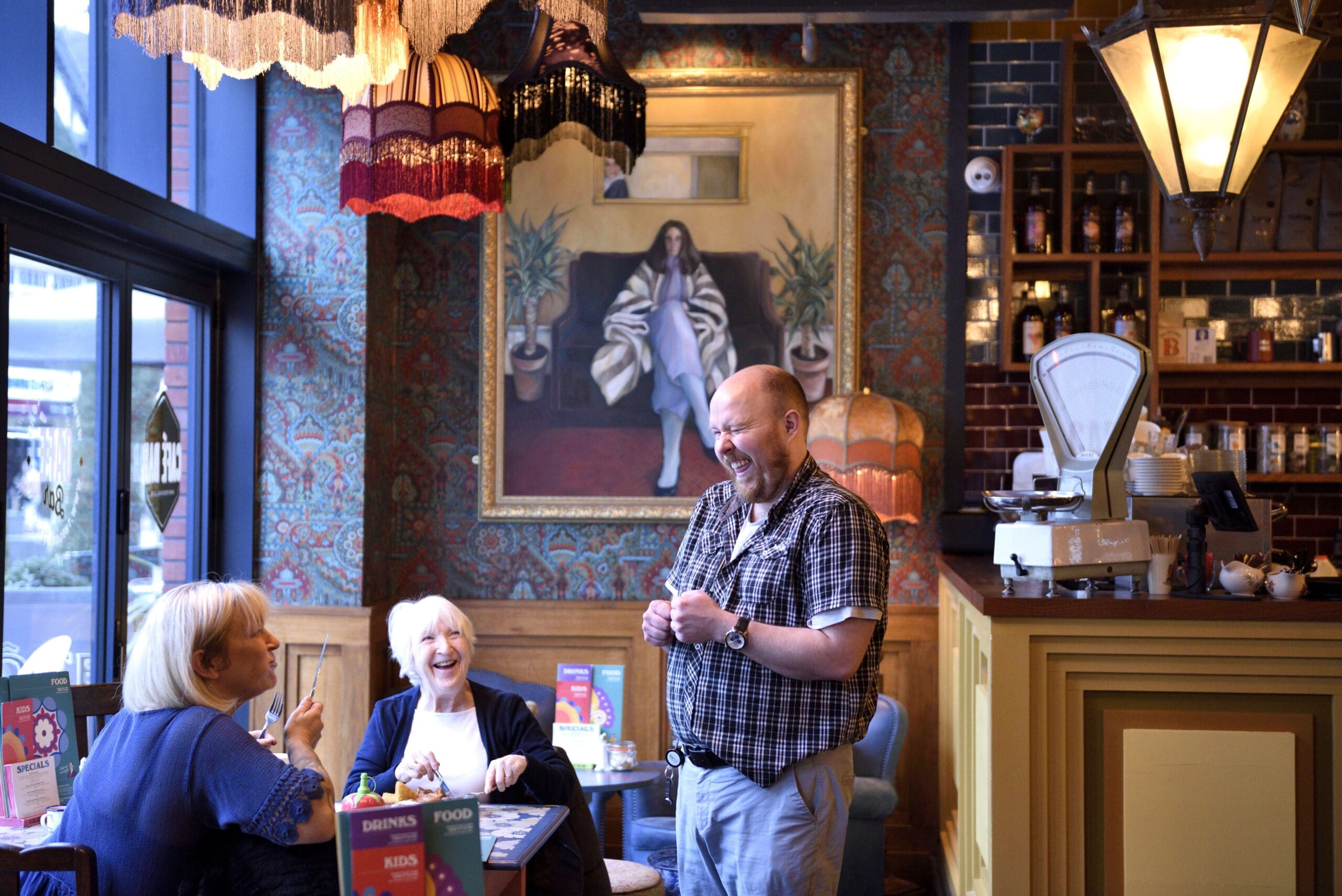 Three people enjoy a lively conversation at a cozy, warmly lit café with vintage decor. A man named Cassio stands smiling and gesturing, while two women laugh. A large portrait and eclectic lamps decorate the background.