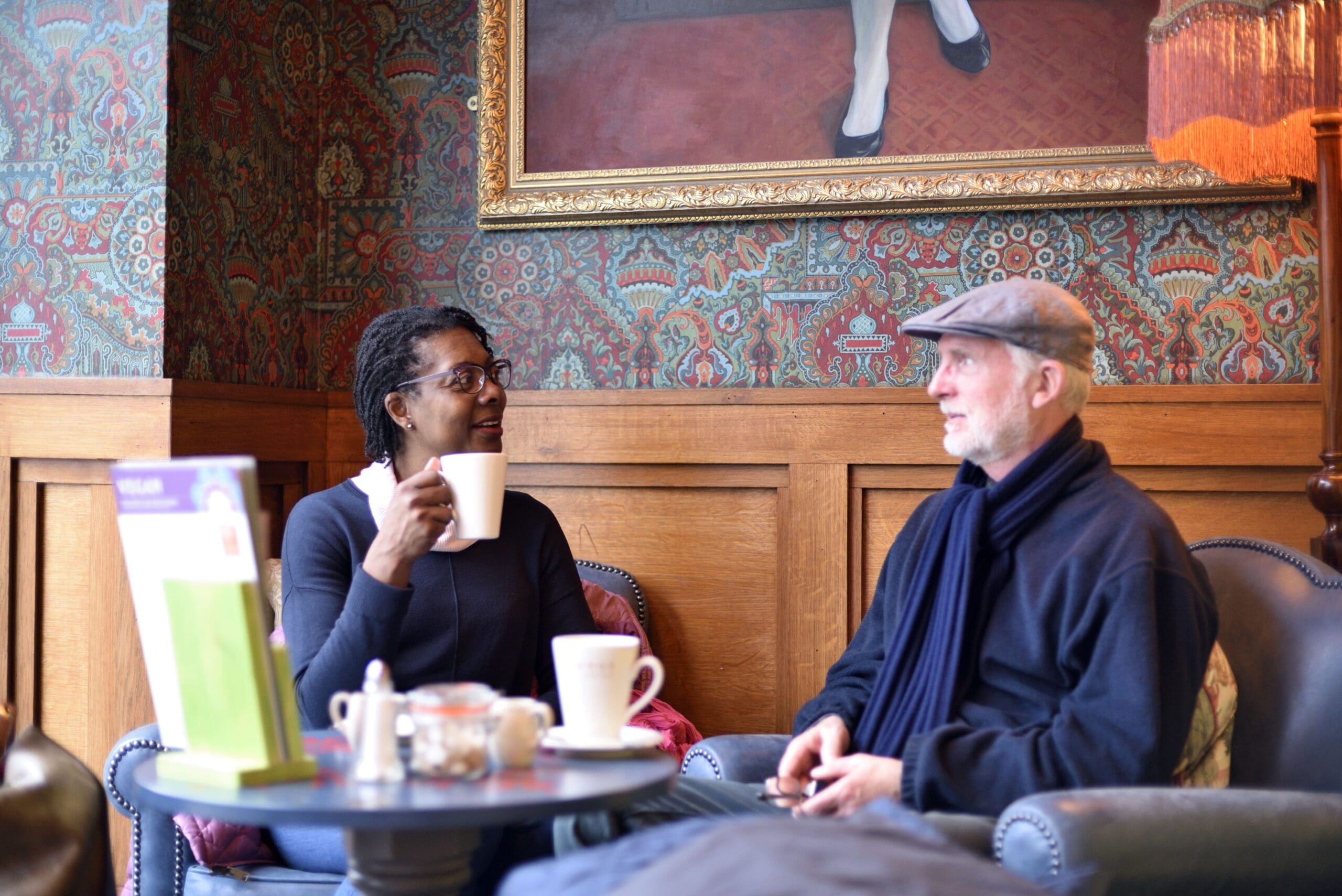 Two people sit at a small round table in the cozy Cassio cafe, having a conversation. One holds a coffee mug, the other wears a scarf and cap. Warm, patterned decor and a large framed painting are visible in the background.