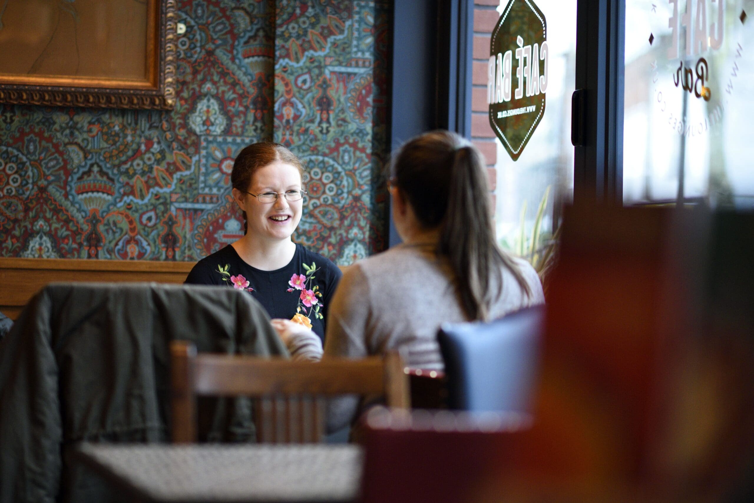 Two women sit at a table in a cozy café, engaged in conversation. Cassio watches as one woman, with red hair and glasses, smiles warmly. The scene is softly lit, with patterned wallpaper and a coat draped over a nearby chair.