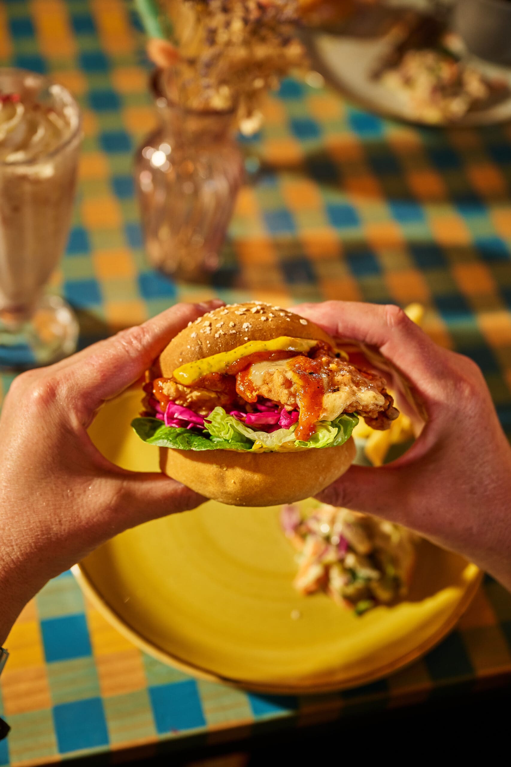 A person holds a chicken burger with lettuce, purple cabbage, onion rings, and sauce on a sesame bun, above a yellow plate on a checkered table, with a milkshake and vase in the background.