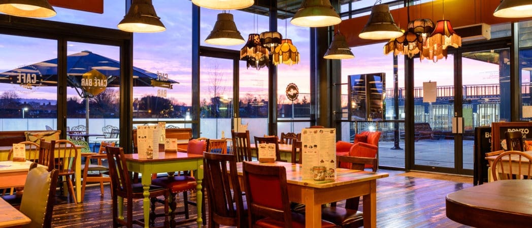 A cozy Bonzo restaurant interior with colorful chairs, wooden tables, and warm hanging lights. Large windows reveal an outdoor patio and a vibrant sunset sky in the background. Menus are placed on each table.