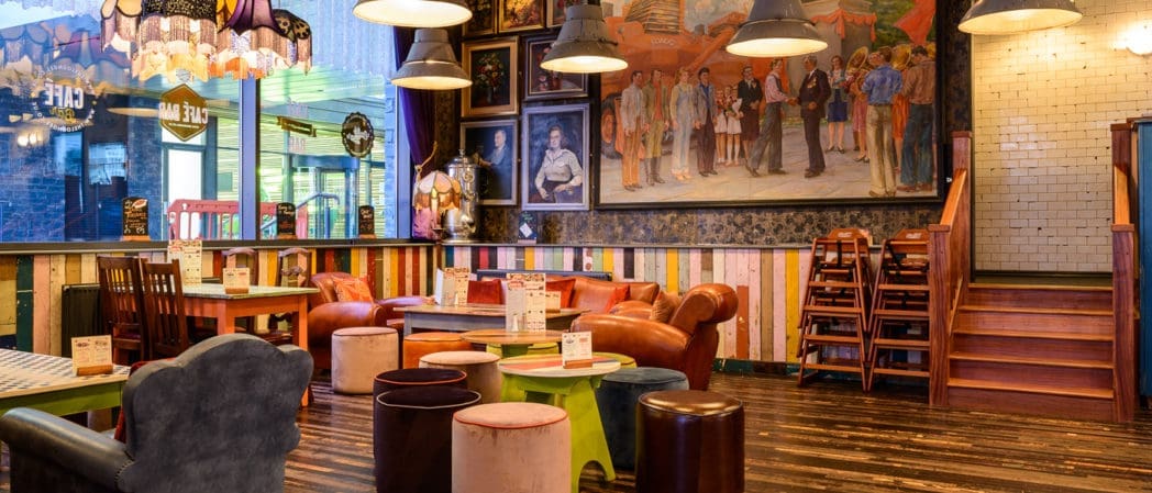 Colorful cafe interior with eclectic furniture, round tables, and mismatched chairs. Bonzo-inspired wall art and paintings add flair. Wooden floors and paneled walls create a cozy vibe, with natural light streaming in from a window on the left.