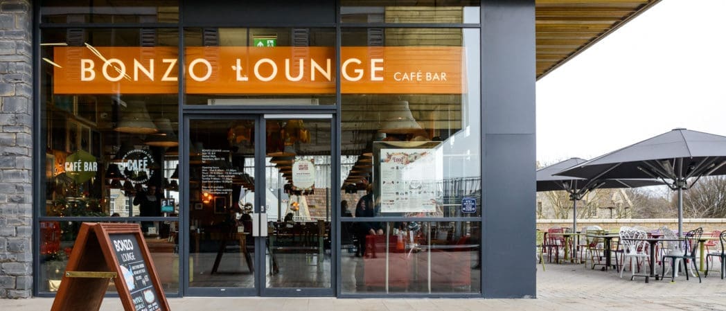 Street view of Bonzo Lounge Café Bar with large windows showing indoor seating, warm lighting, and a Bonzo-branded sign above the entrance. Outdoor seating with tables, chairs, and umbrellas is visible to the right.