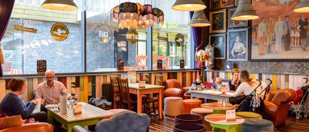 A colorful, eclectic café with mismatched furniture—Bonzo’s signature style—welcomes families and friends dining at tables. Large windows let in natural light, while walls decorated with portraits and artwork add to the lively atmosphere.