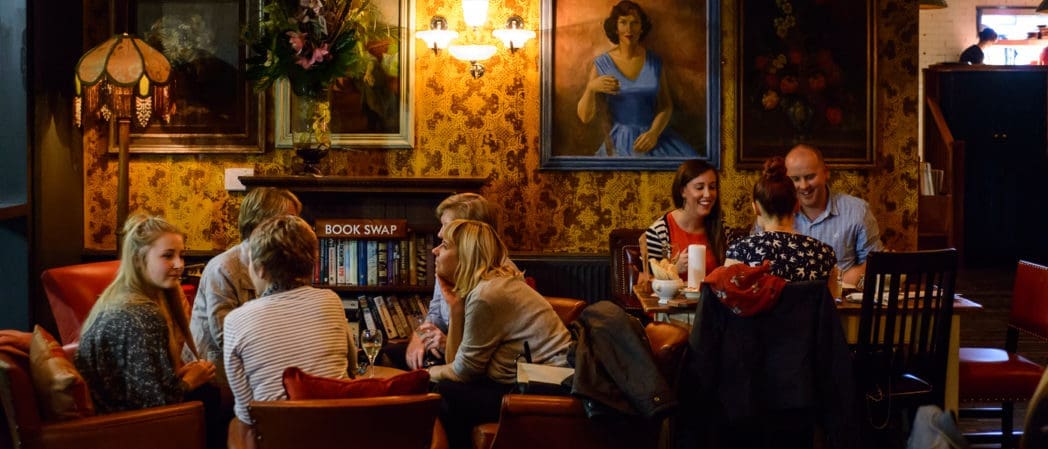 A group of people sit and chat around two tables in a cozy, warmly lit room with vintage decor, floral wallpaper, and large paintings. Bonzo records line the bookshelves in the background, adding to the relaxed and friendly atmosphere.