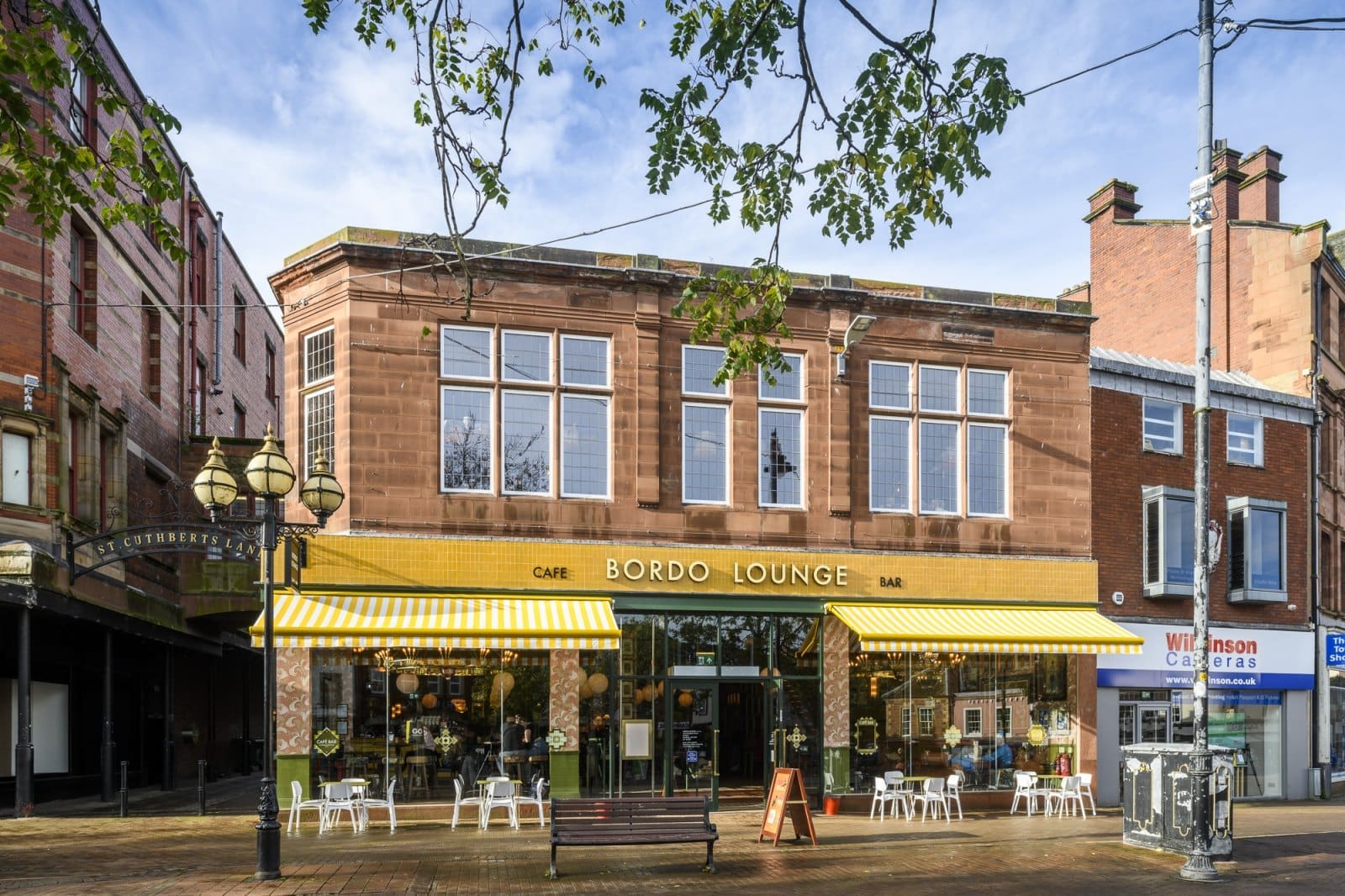 The image shows Bordo Lounge, a vibrant café and bar housed in a two-story brick building with large windows and yellow-striped awnings. Outdoor tables and chairs are set up on the pavement, while a tree branch artfully frames the Bordo scene.