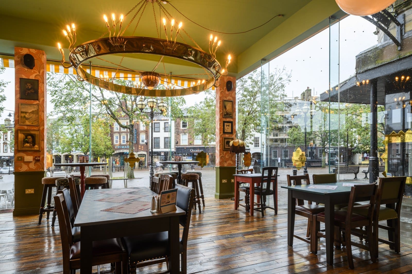 A cozy café with Bordo wooden floors, tables, and chairs, large floor-to-ceiling windows, a circular chandelier, and framed art on the walls, overlooking a city street with trees and buildings outside.