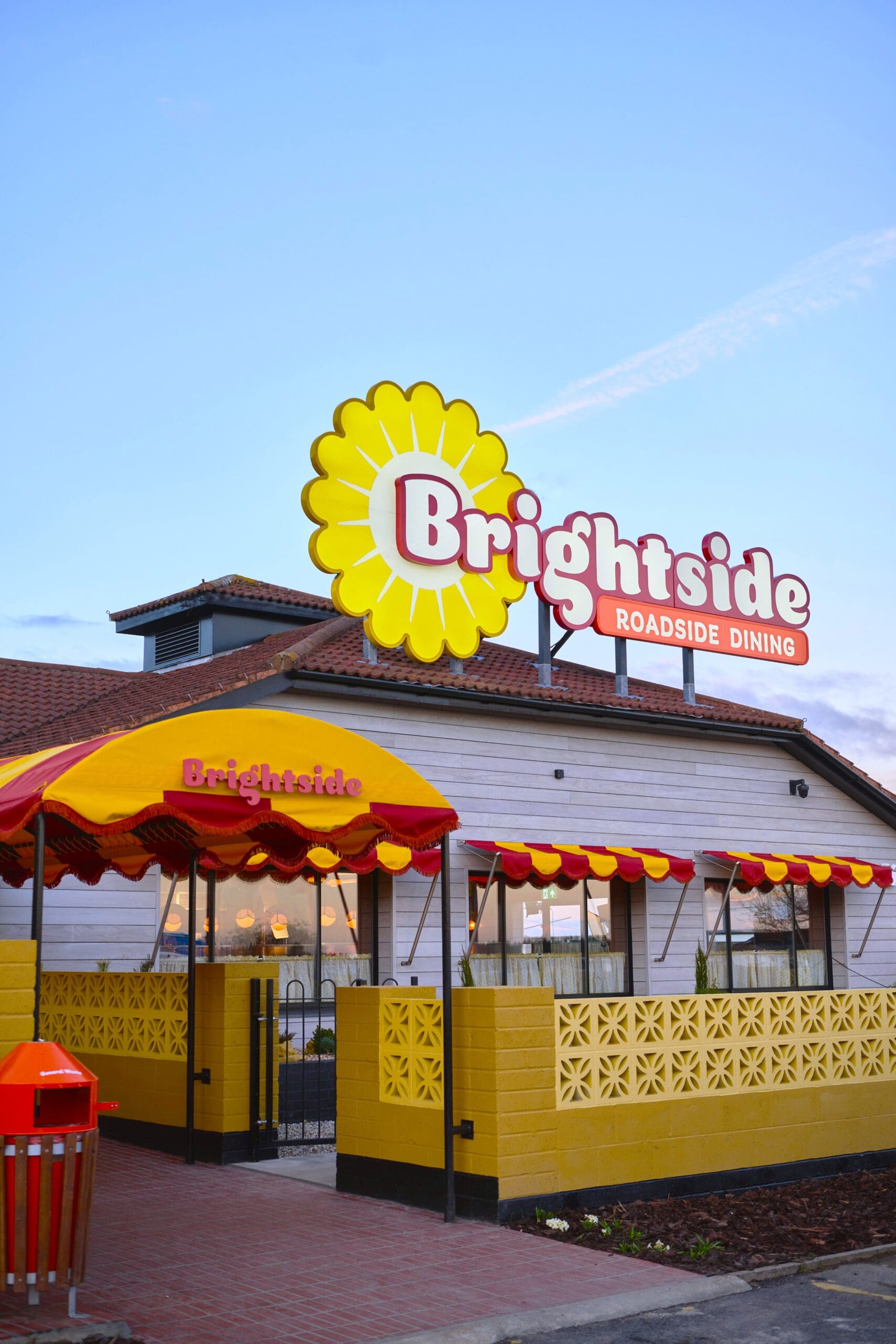 A roadside diner named Brightside with a large yellow flower logo, red and yellow awning, and outdoor seating area, photographed at dusk.
